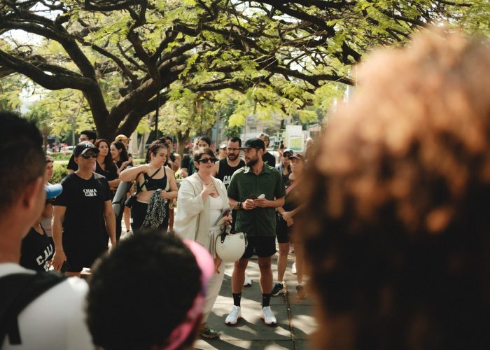 Na foto vemos uma das fundadoras do VerboGentileza, Pat Tavares, vestida de blusa e calça branca e usando óculos escuros. Ela está ao centro de uma roda de variadas pessoas que participaram de uma caminhada, portanto usam roupas para prática de exercício. Todos estão debaixo de árvores em um espaço público.
