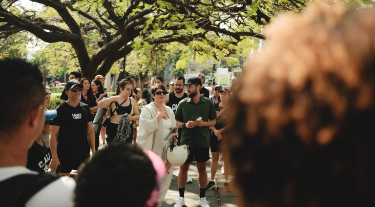 Na foto vemos uma das fundadoras do VerboGentileza, Pat Tavares, vestida de blusa e calça branca e usando óculos escuros. Ela está ao centro de uma roda de variadas pessoas que participaram de uma caminhada, portanto usam roupas para prática de exercício. Todos estão debaixo de árvores em um espaço público.